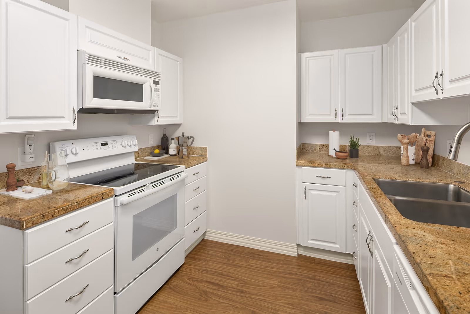 A clean and modern kitchen with white cabinets, granite countertops, a white electric stove with an oven, a microwave above the stove, a double stainless steel sink, and wooden flooring. Various kitchen utensils and small decorative items are placed on the countertops.