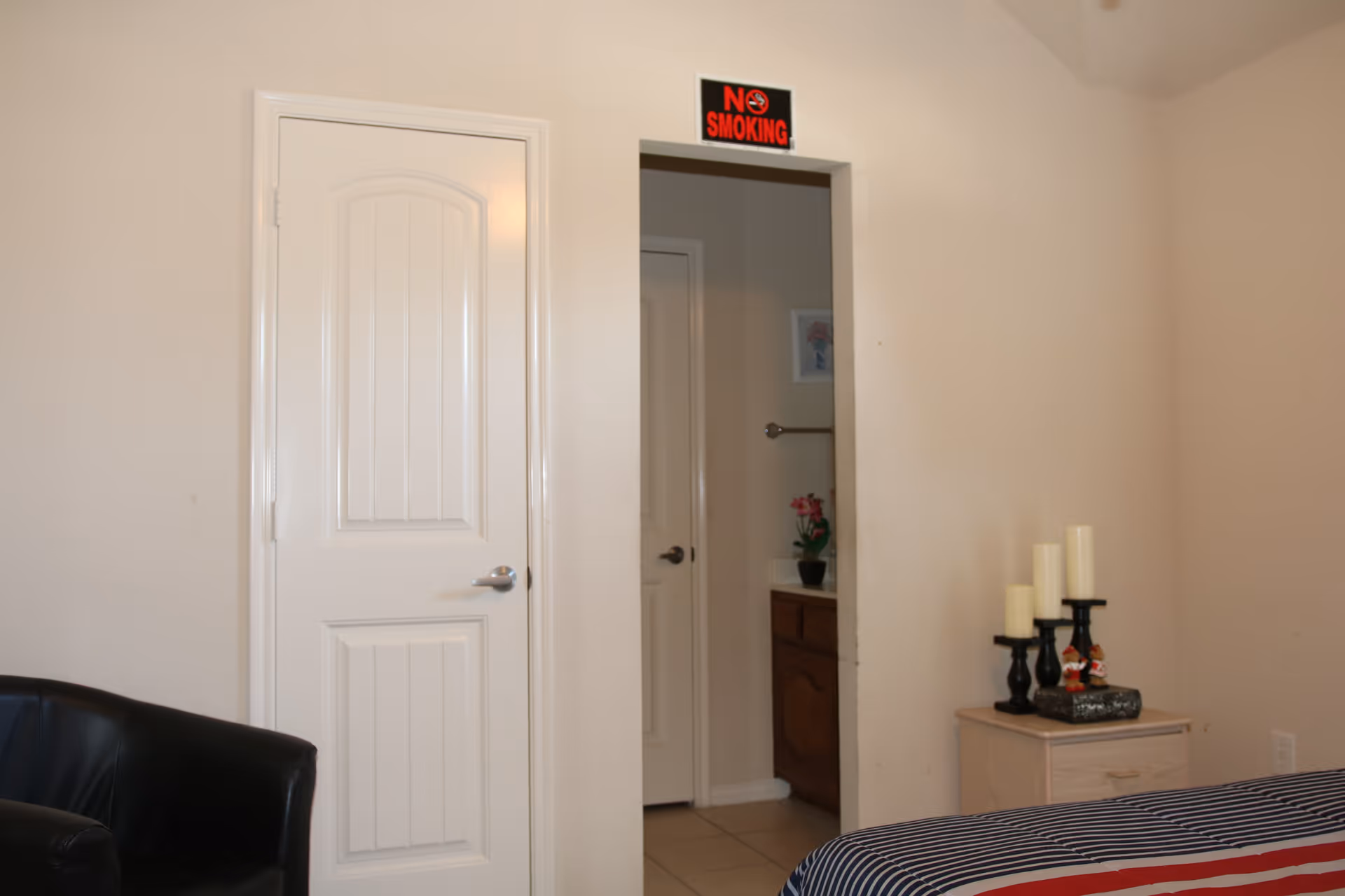 Bedroom interior showing a closed white door and an open doorway with a "NO SMOKING" sign, a striped bed, small nightstand with candles, and a black chair.