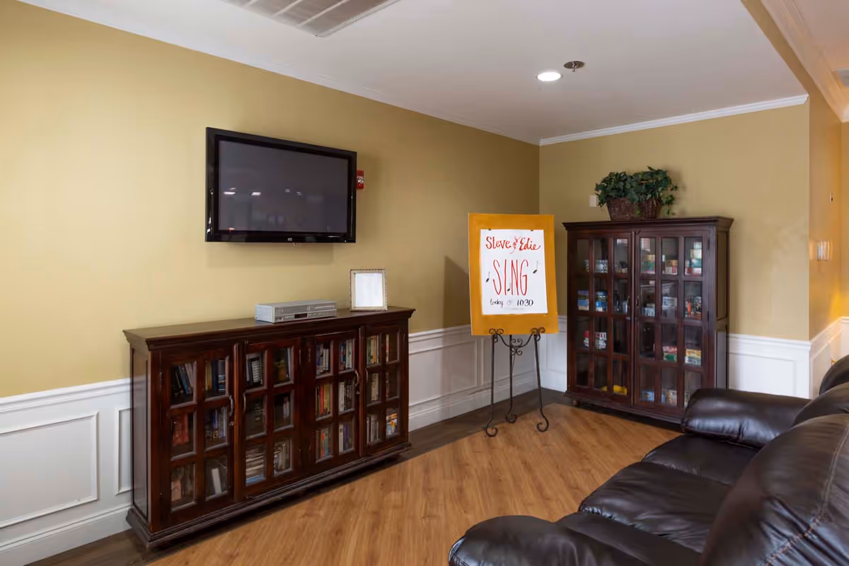 A cozy living room area with a wall-mounted flat screen TV above a wooden cabinet filled with books. Next to it is a wooden display cabinet with glass doors containing various items. A sign on an easel reads 'Steve & Edie SING Looking @ 10:30'. A dark leather couch is partially visible on the right side. The room has light yellow walls with white wainscoting and wood flooring.