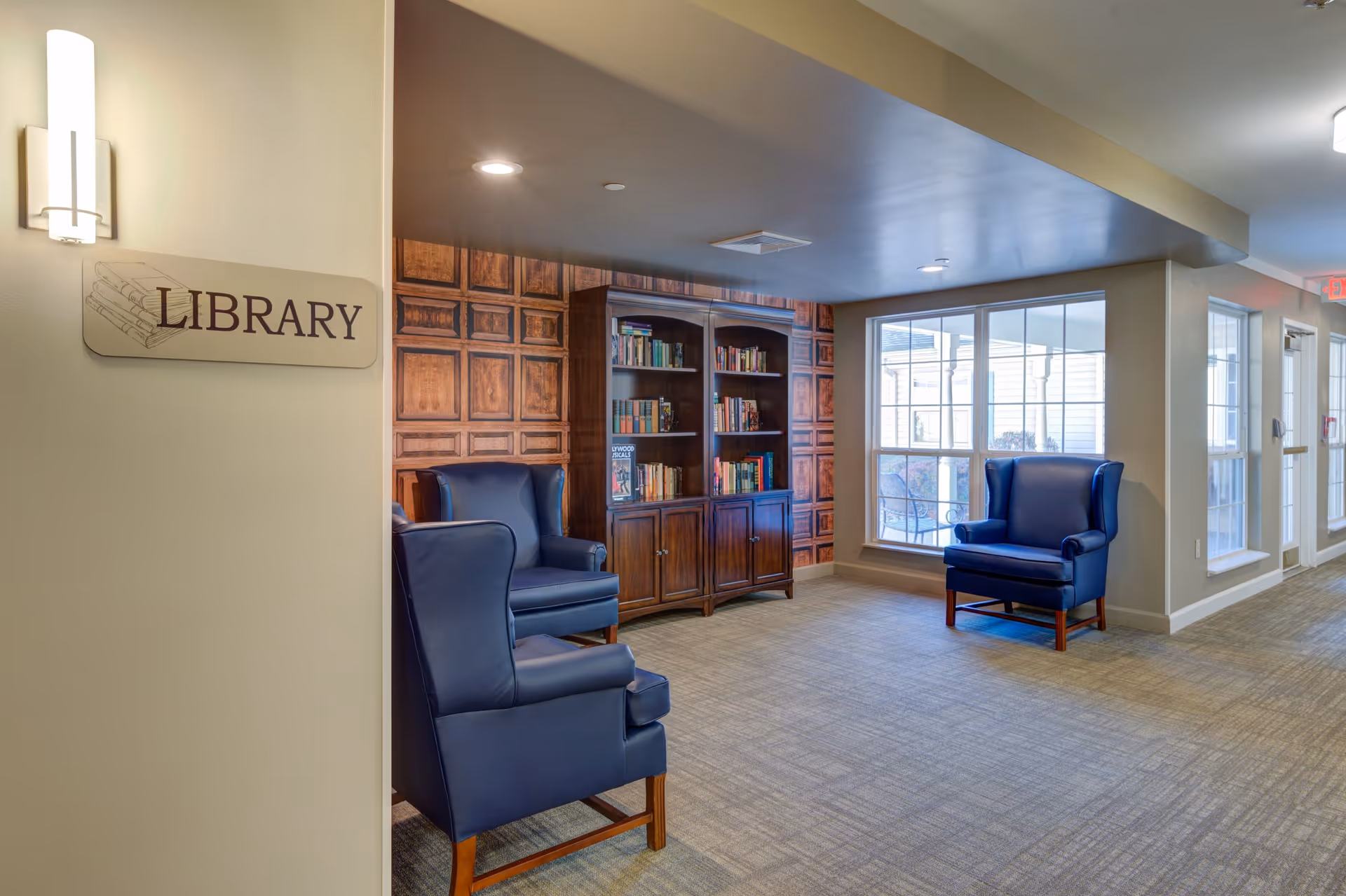 Interior view of a library area in a senior living facility with a wooden bookshelf filled with books, two blue upholstered armchairs, large windows letting in natural light, and a sign on the wall that reads 'LIBRARY'.