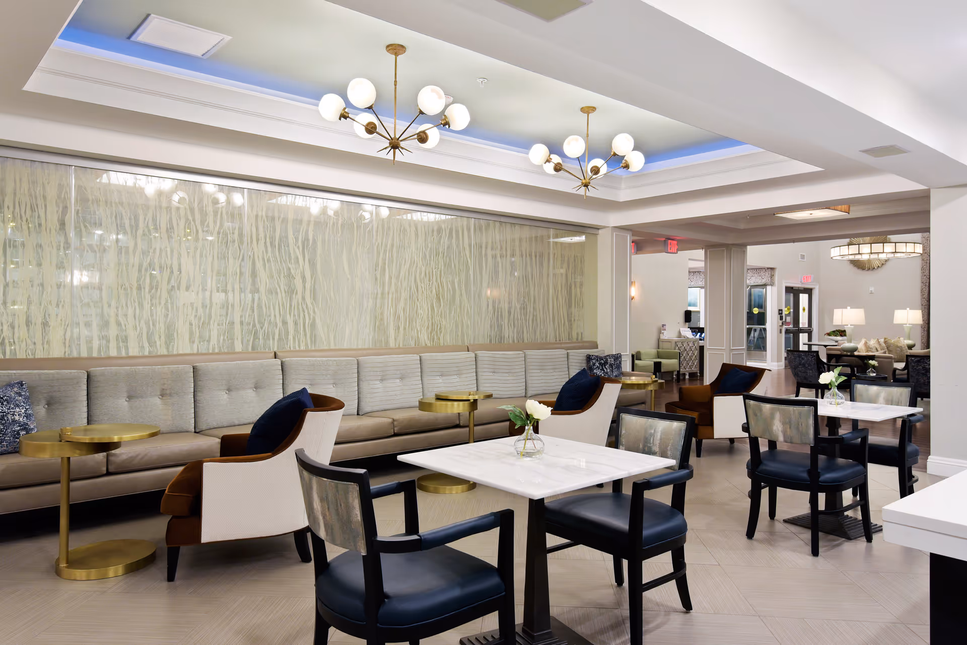A spacious and modern common area in a senior living facility featuring a long beige tufted sofa along the wall with several small round gold side tables. There are multiple seating arrangements including armchairs with dark cushions and black chairs around white marble-top tables. The ceiling has recessed lighting and two stylish chandeliers with globe bulbs. The room has a neutral color palette with light walls and flooring, and decorative elements like a textured wall panel and fresh flowers on the tables.