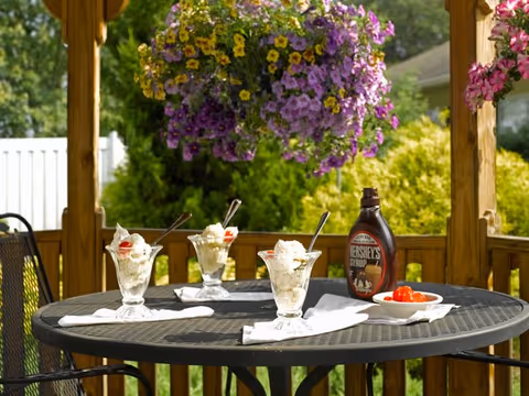 Round outdoor patio table on a wooden deck set with three ice cream sundaes, a bottle of Hershey's syrup, and a small bowl of cherries under hanging purple flowers.