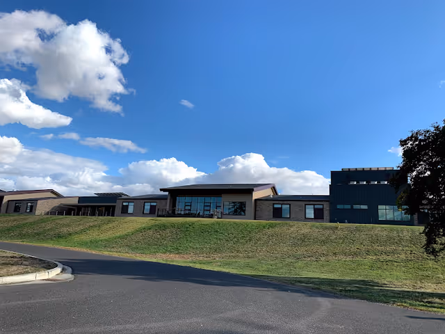 Exterior view of a modern single-story building with large windows, situated on a grassy hill under a blue sky with scattered clouds. A paved road curves in front of the building, and a tree is partially visible on the right side.