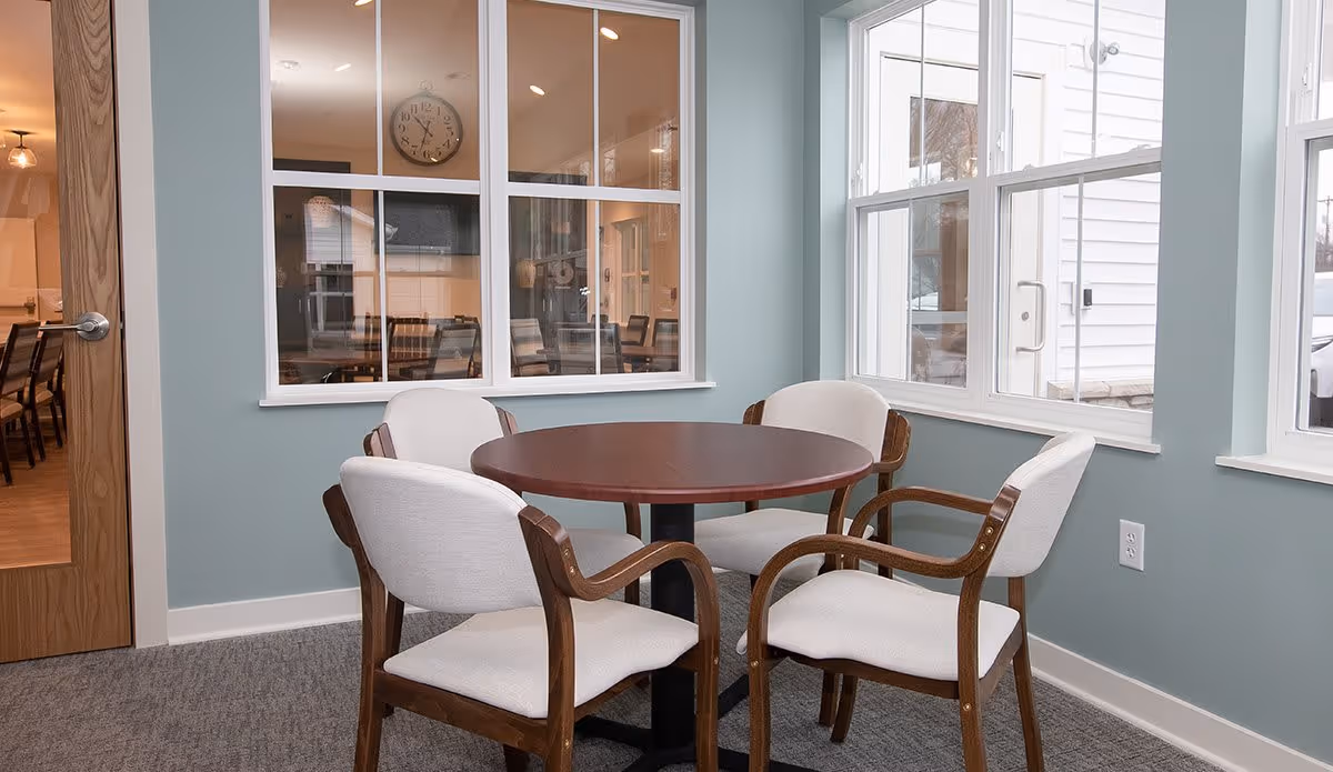 A small seating area with a round wooden table and four cushioned chairs with wooden arms, situated next to large windows letting in natural light. Through an interior window, a dining area with tables and chairs and a wall clock is visible.