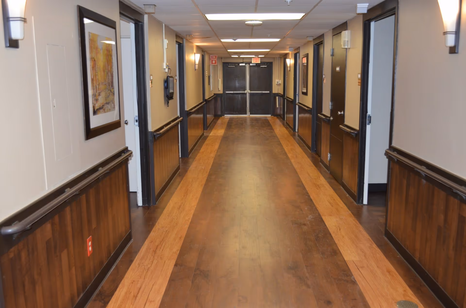 Well-lit interior hallway of a nursing facility with wood floors, handrails, framed art, multiple doors, and double exit doors at the far end.