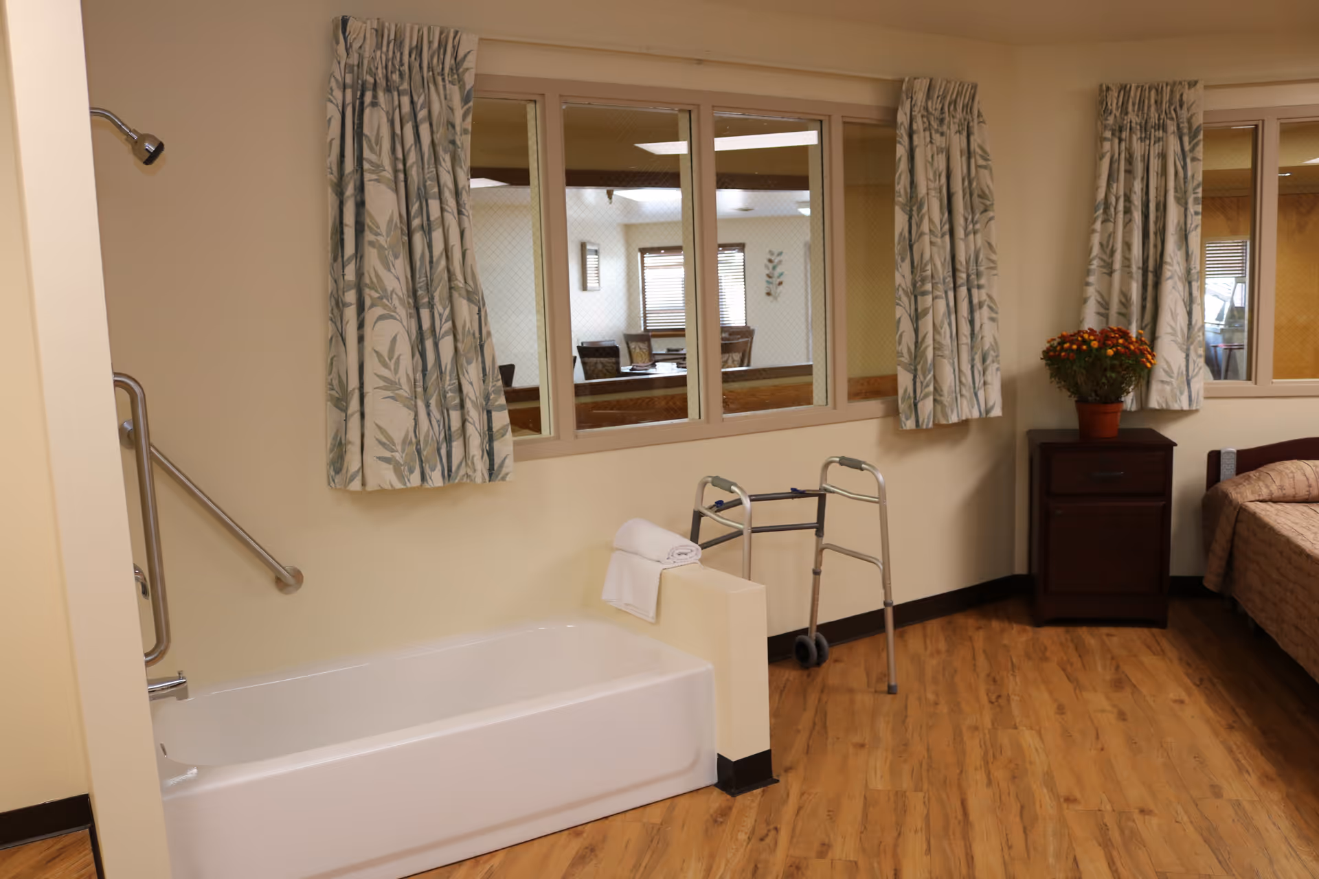 A senior living facility room featuring a white bathtub with a grab bar, a walker, a wooden nightstand with a potted plant, and a bed partially visible on the right. The room has wood flooring, beige walls, and windows with floral curtains looking into another room with tables and chairs.