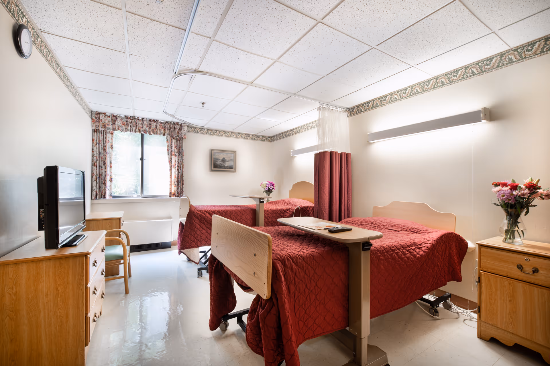 A bright room with two hospital-style beds covered in red quilts, each with an overbed table. There is a wooden dresser with a TV on top, a wooden nightstand with a vase of flowers, a chair, a window with floral curtains, and a wall clock. The room has a ceiling track for privacy curtains and fluorescent lighting.