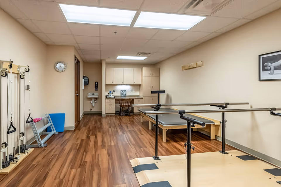 A rehabilitation or physical therapy room with wooden flooring, parallel bars for walking exercises, exercise equipment on the left wall, a desk with a chair and cabinets in the back, and a black and white framed photo of a baby on the right wall.