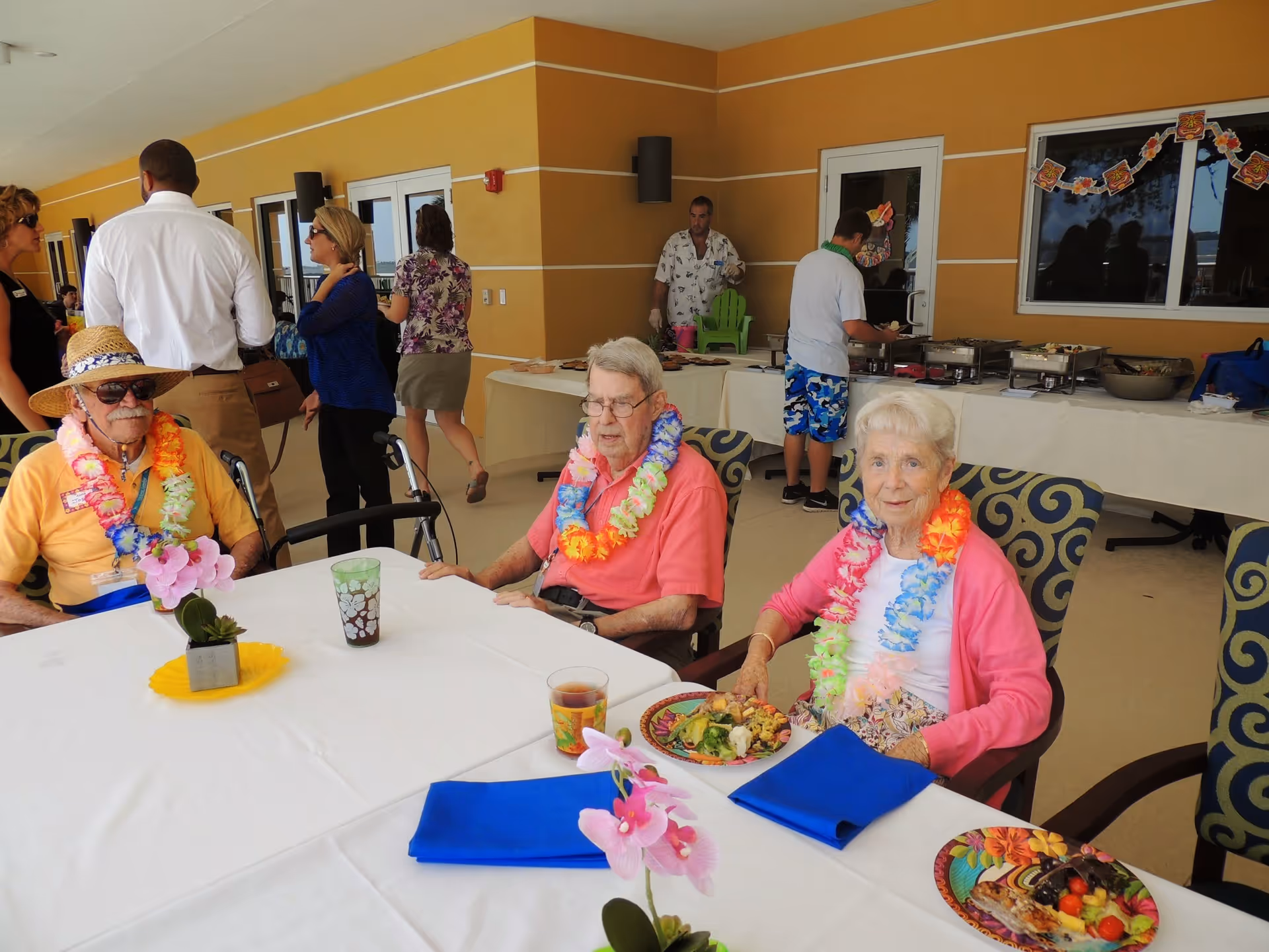 Three elderly people wearing colorful leis sit at a table with plates of food and drinks in front of them. Behind them, several people are standing and serving themselves food from a buffet table set up against a yellow wall under a covered outdoor area.