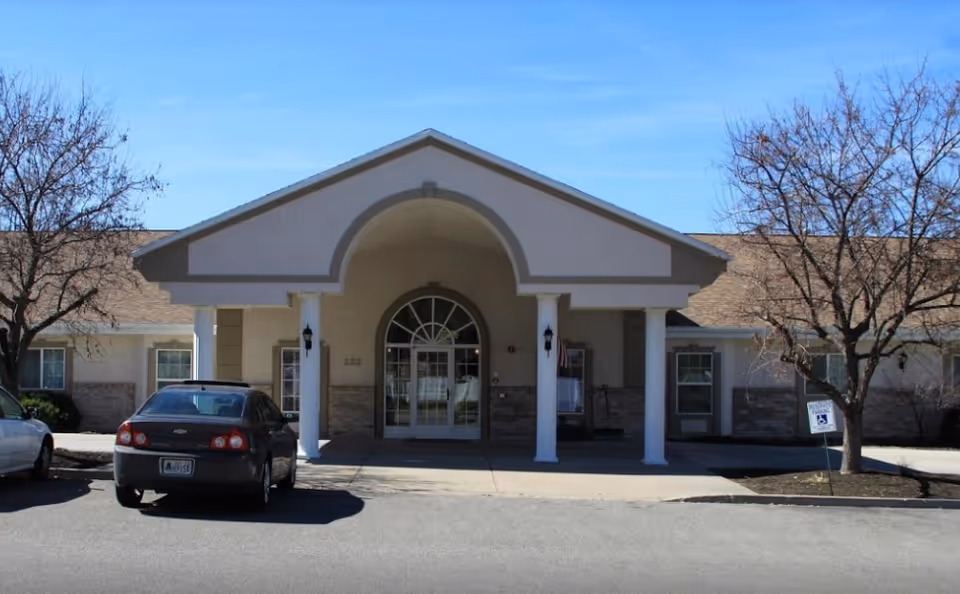 Front exterior view of Cache Valley Assisted Living and Memory Care building with a covered entrance supported by white columns, two parked cars in front, leafless trees on either side, and a clear blue sky.