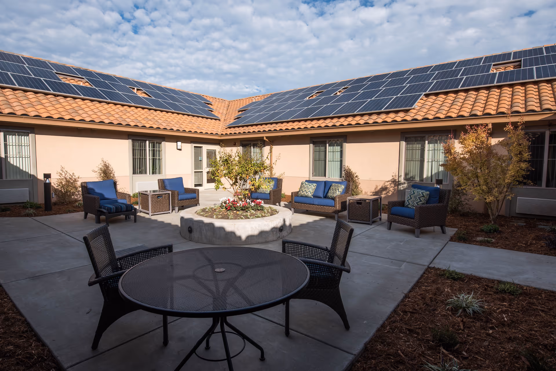 Outdoor courtyard area with patio furniture including a round metal table with four chairs in the foreground and several cushioned chairs and sofas arranged around a circular raised planter with a small tree and flowers. The building surrounding the courtyard has a tiled roof with solar panels and beige walls with windows.