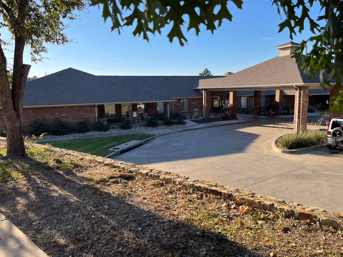 Front entrance and circular driveway of a single-story brick senior living building framed by trees and landscaping.