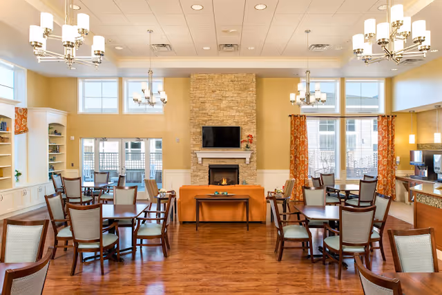 A spacious and well-lit dining room with multiple wooden tables and chairs arranged neatly. The room features a stone fireplace with a mounted flat-screen TV above it, orange patterned curtains on the windows, and modern chandeliers hanging from the ceiling. There is a coffee station on the right side and large windows allowing natural light to fill the space.