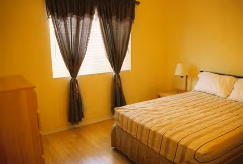 Yellow bedroom with striped bed, dresser, nightstand, and window with dark curtains.