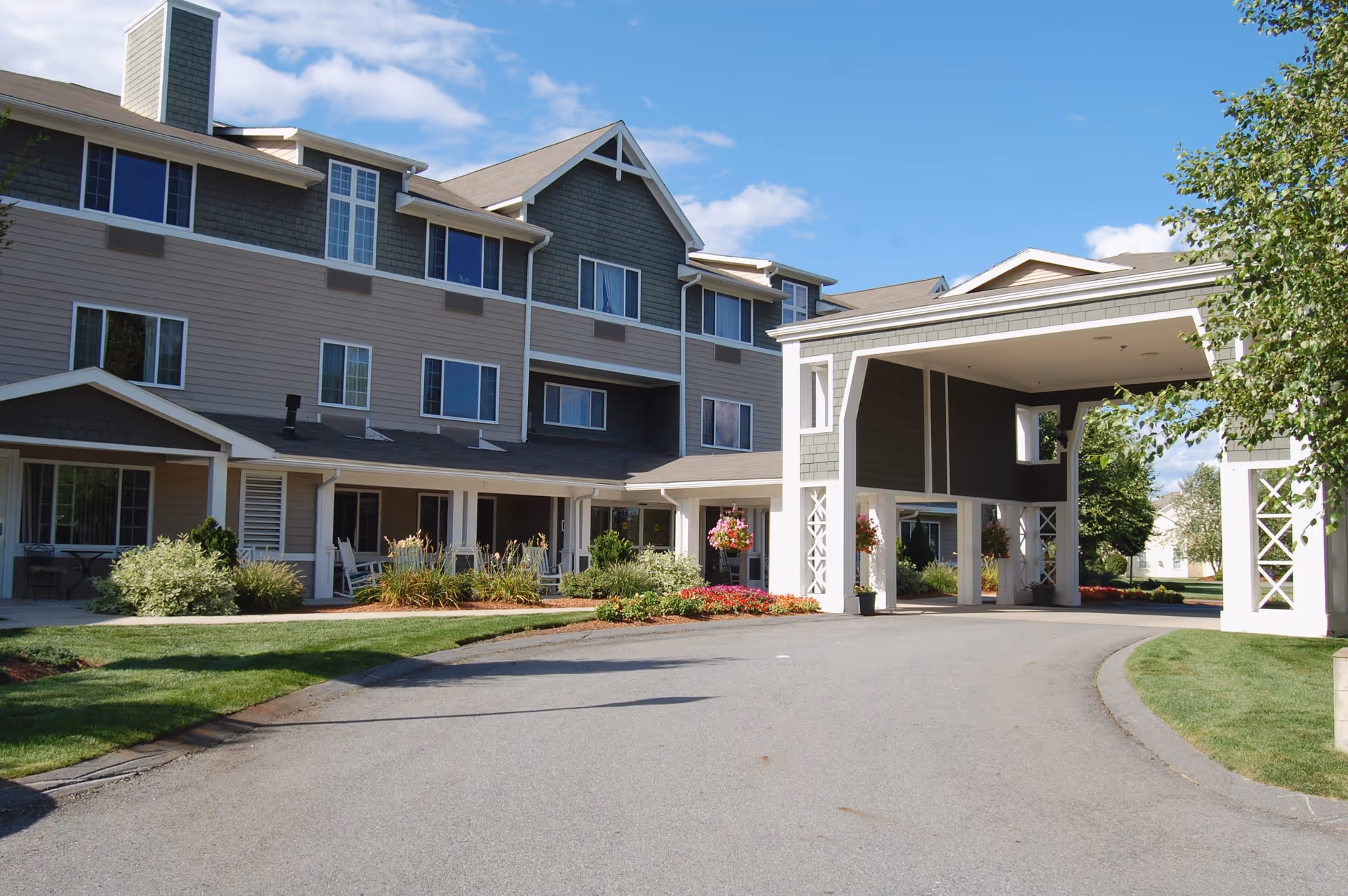 Exterior view of Bayberry at Emerald Court senior living facility showing a multi-story building with gray and beige siding, multiple windows, a covered entrance with white pillars, landscaped greenery, and a clear blue sky.