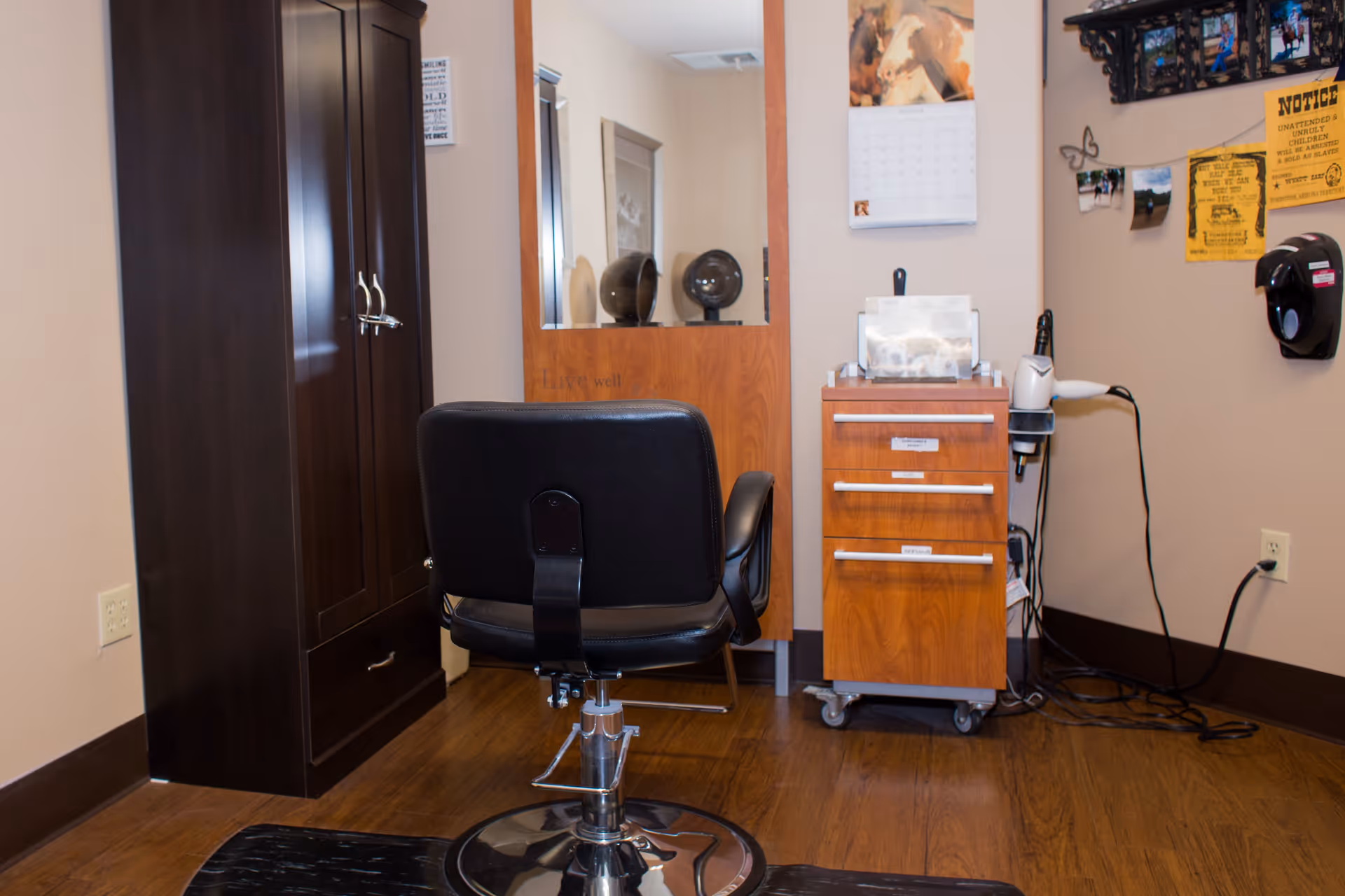 Interior view of a small salon or barber area with a black salon chair facing a mirror mounted on a wooden cabinet. To the right, there is a wooden rolling cart with drawers, a hairdryer, and various hair care items. The walls have a calendar, notices, and a mounted soap or sanitizer dispenser.