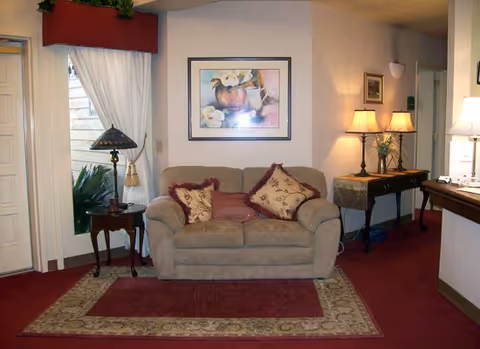 A cozy living room area with a beige loveseat adorned with decorative pillows. Behind the loveseat is a framed painting of flowers. To the left, there is a window with white curtains and a small wooden side table with a decorative lamp. To the right, there is a console table with two lamps and some decorative items. The floor is carpeted with a patterned area rug and a smaller red rug on top.