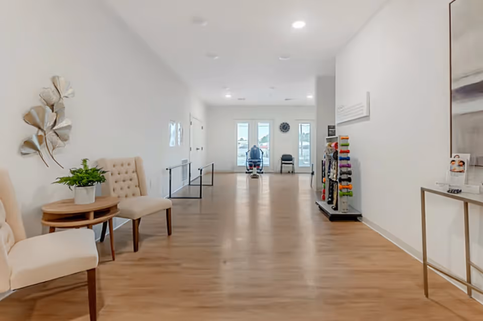 A bright, spacious hallway with light wooden flooring and white walls. On the left side, there are two beige upholstered chairs with a small round wooden table holding a potted plant. On the right side, there is a rack with colorful dumbbells and a narrow console table with brochures and a decorative item. At the far end, a person is seated on a mobility scooter near large windows letting in natural light.