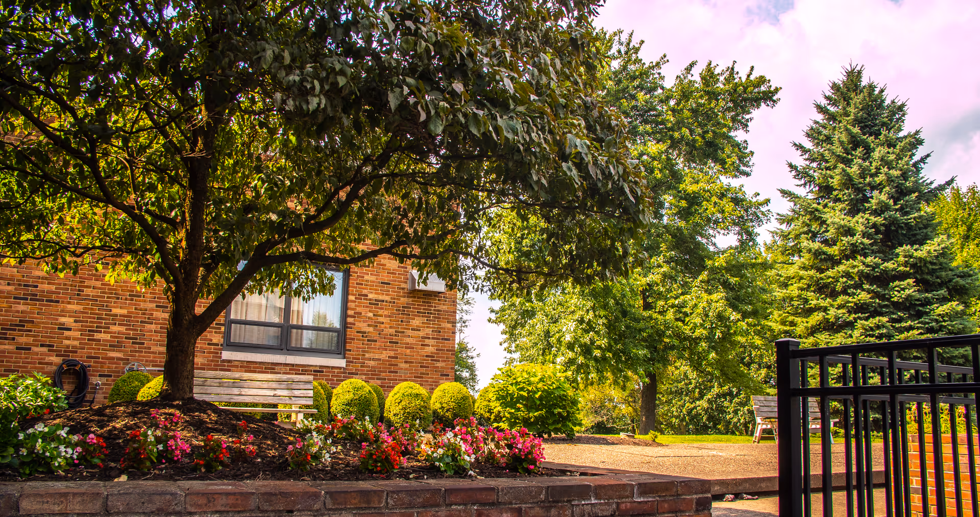 Brick building exterior with a shaded bench, landscaped flowerbeds and trees in a courtyard.