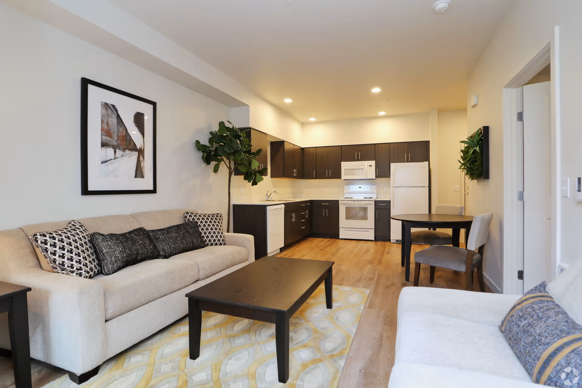 A modern open-concept living space featuring a beige sofa with patterned cushions, a dark wooden coffee table on a light patterned rug, and a kitchen area with dark cabinets, white appliances including a refrigerator, stove, microwave, and dishwasher. There is a small round dining table with two chairs, a framed artwork on the wall, and a tall green plant in the corner.