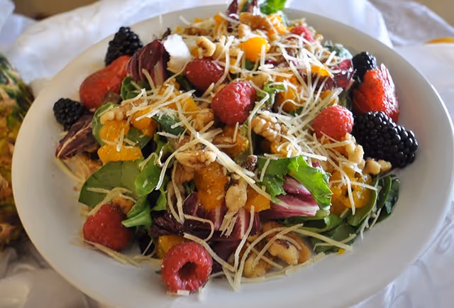 A close-up of a fresh salad in a white bowl containing mixed greens, raspberries, blackberries, mandarin orange segments, walnuts, and shredded cheese.