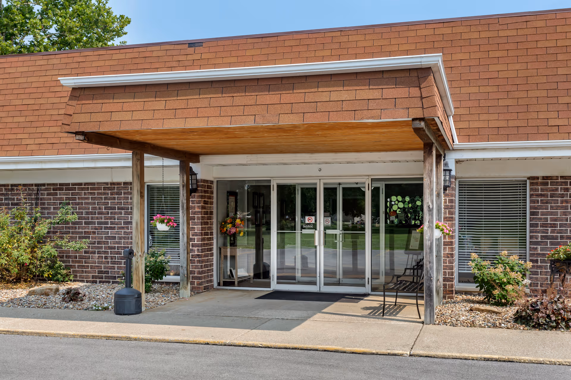 Covered wooden awning over the glass double-door entrance of a brick building with potted flowers and landscaping.