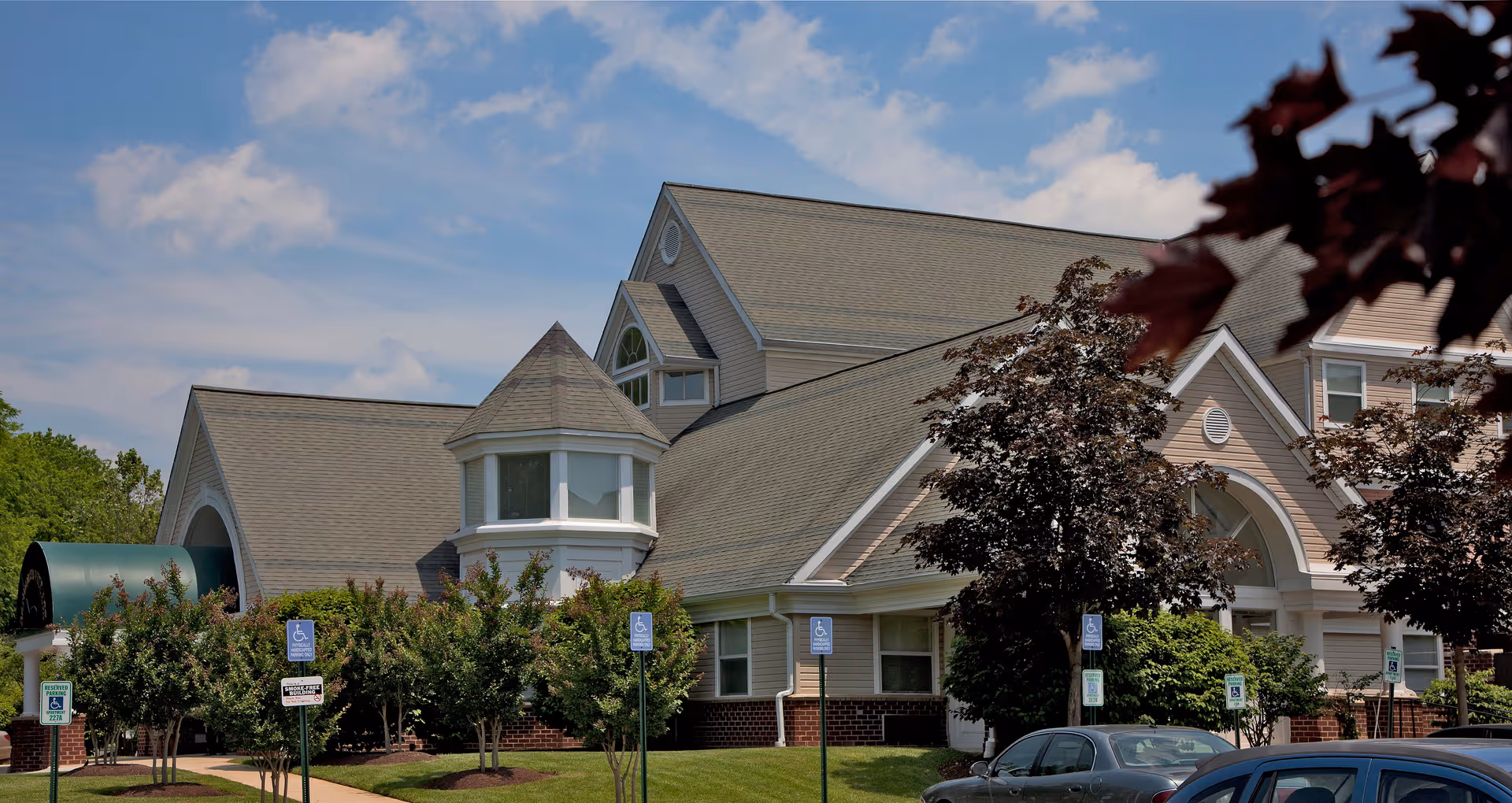 Front exterior of a multi-gabled residential building with trees, accessible parking signs, and parked cars.