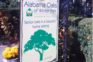 Sign for Alabama Oaks of Winter Park Assisted Living, featuring a green tree graphic and text that reads 'Senior care in a beautiful home setting', surrounded by plants and flowers outdoors.