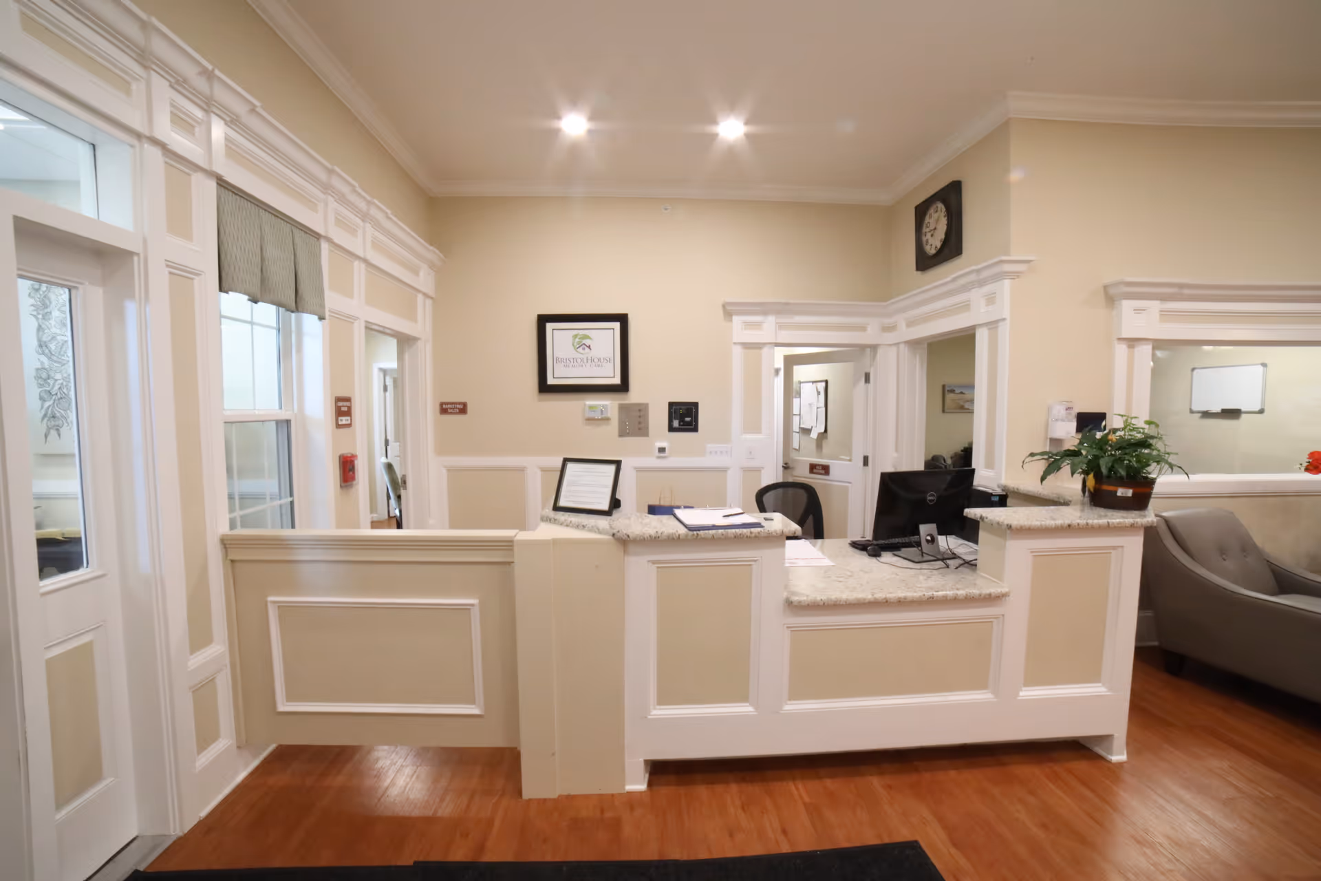 Reception desk area inside Bristol House Memory Care facility with a computer, office chair, potted plant, and framed signage on the wall. The space has beige walls, white trim, wooden flooring, and a clock mounted above the desk.