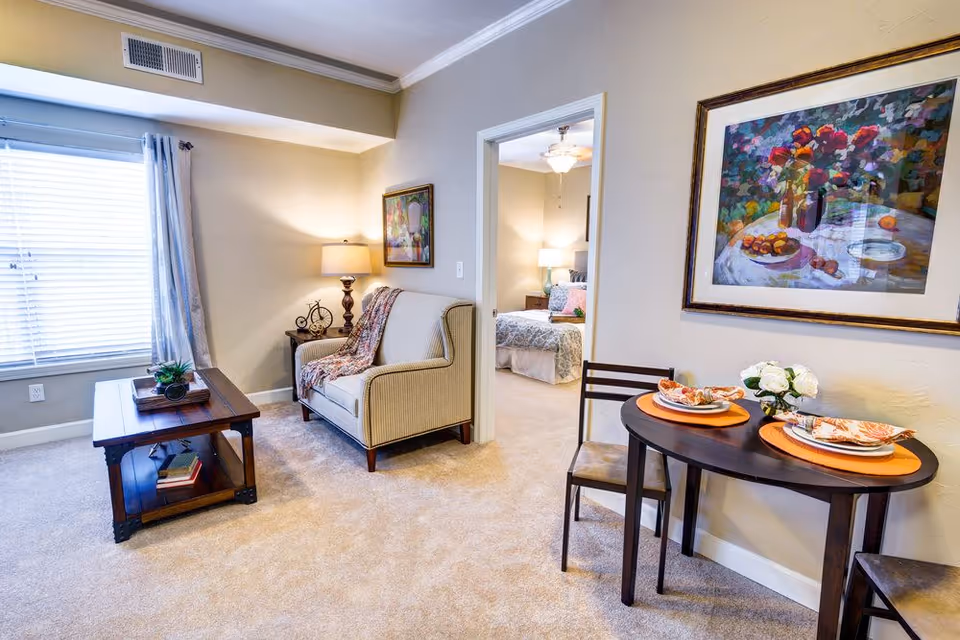 Cozy living room with an armchair, coffee table, small dining table set for two, and a view into a bedroom.