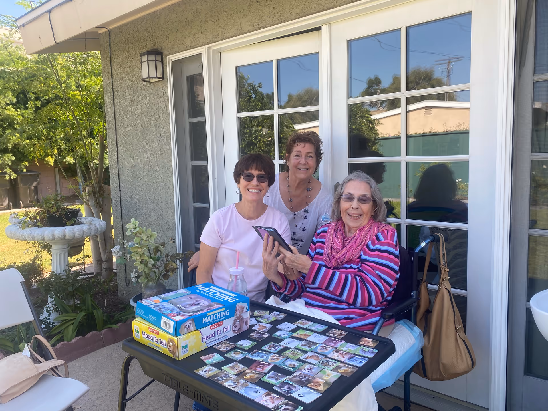 Three elderly women sitting outside a senior wellness home, smiling and playing a matching card game on a table. One woman is in a wheelchair wearing a striped sweater and pink scarf, while the other two women are seated and standing beside her. They are in a garden area with greenery and a birdbath visible in the background.
