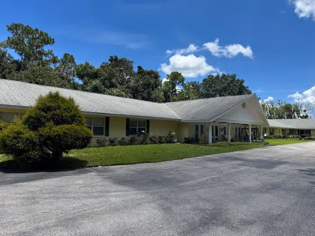 Single-story yellow retirement center building with a covered entrance, lawn, shrubs, and a paved driveway under a blue sky.