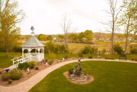 A peaceful outdoor garden area featuring a white gazebo with a small weather vane on top, surrounded by a curved brick pathway and landscaped with bushes and small statues. There is a green lawn with a decorative sculpture in the center and trees in the background under a cloudy sky.