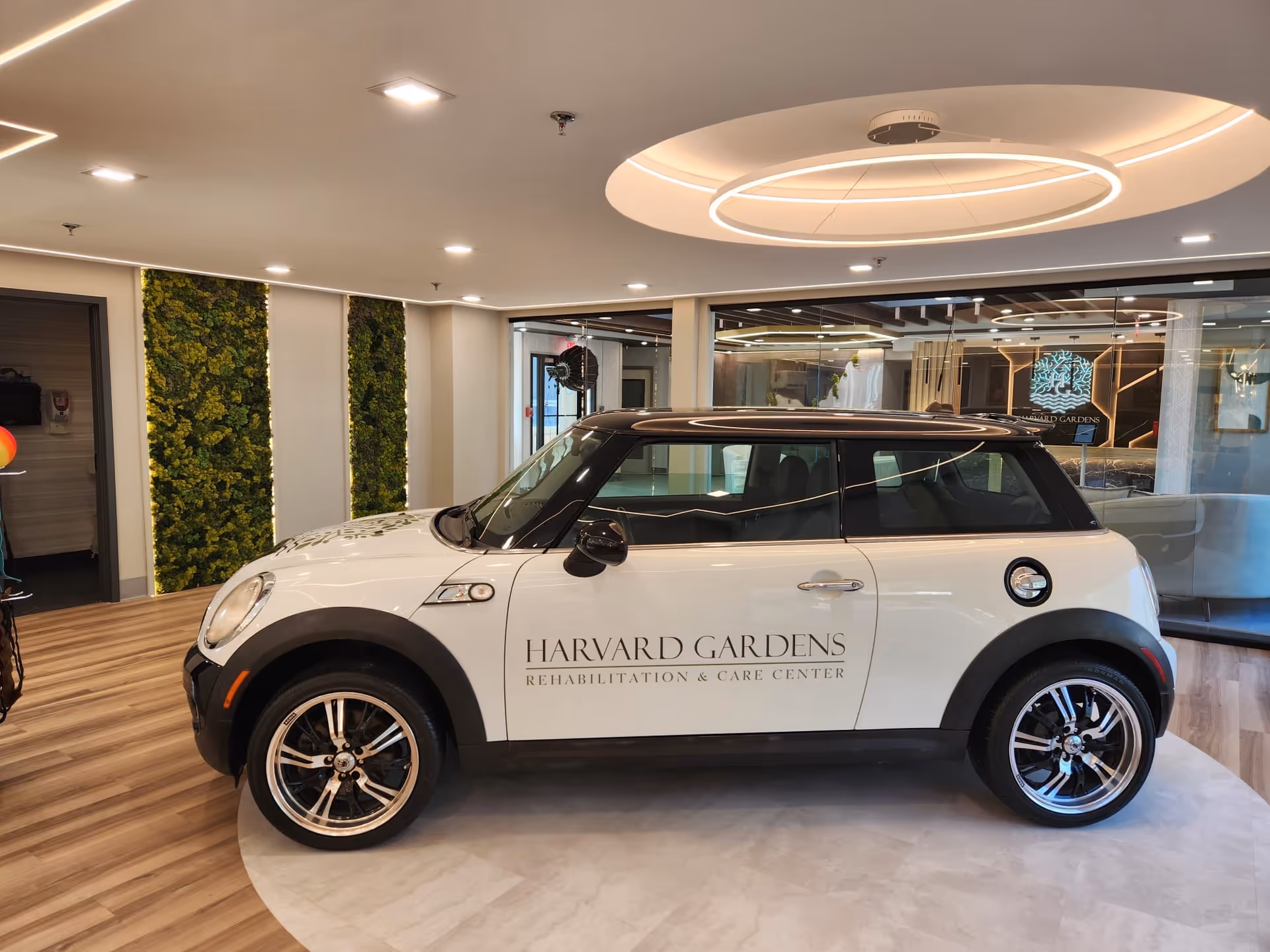 A white and black Mini Cooper car with the text 'Harvard Gardens Rehabilitation & Care Center' on its side is displayed indoors on a circular platform. The room has modern lighting, wood flooring, and green vertical plant panels on the walls. Glass walls and a seating area are visible in the background.