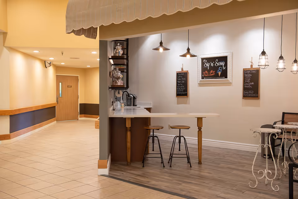 A cozy indoor cafe area with a counter and two wooden stools, pendant lights hanging from the ceiling, and chalkboard menus on the wall. The counter area has jars and a coffee machine. Adjacent to the cafe is a hallway with beige walls, tiled floor, and a wooden door at the end.
