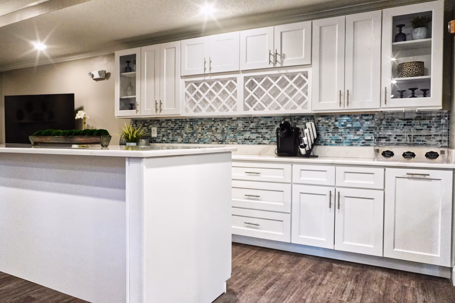 Modern kitchen area with white cabinets, a mosaic tile backsplash, a coffee maker, and a white island countertop. The kitchen has wooden flooring and some decorative plants and items on the counter and shelves.