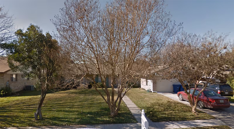 Front view of a single-story suburban house with leafless trees on a lawn, a driveway, and parked cars.