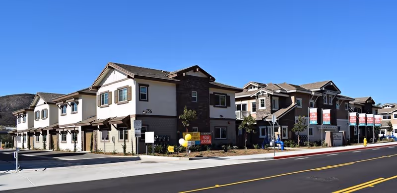 Exterior view of a modern multi-unit residential building with beige and brown tones, featuring multiple windows, balconies, and a well-maintained sidewalk. Several signs and balloons indicate that leasing is now available. The sky is clear and blue.