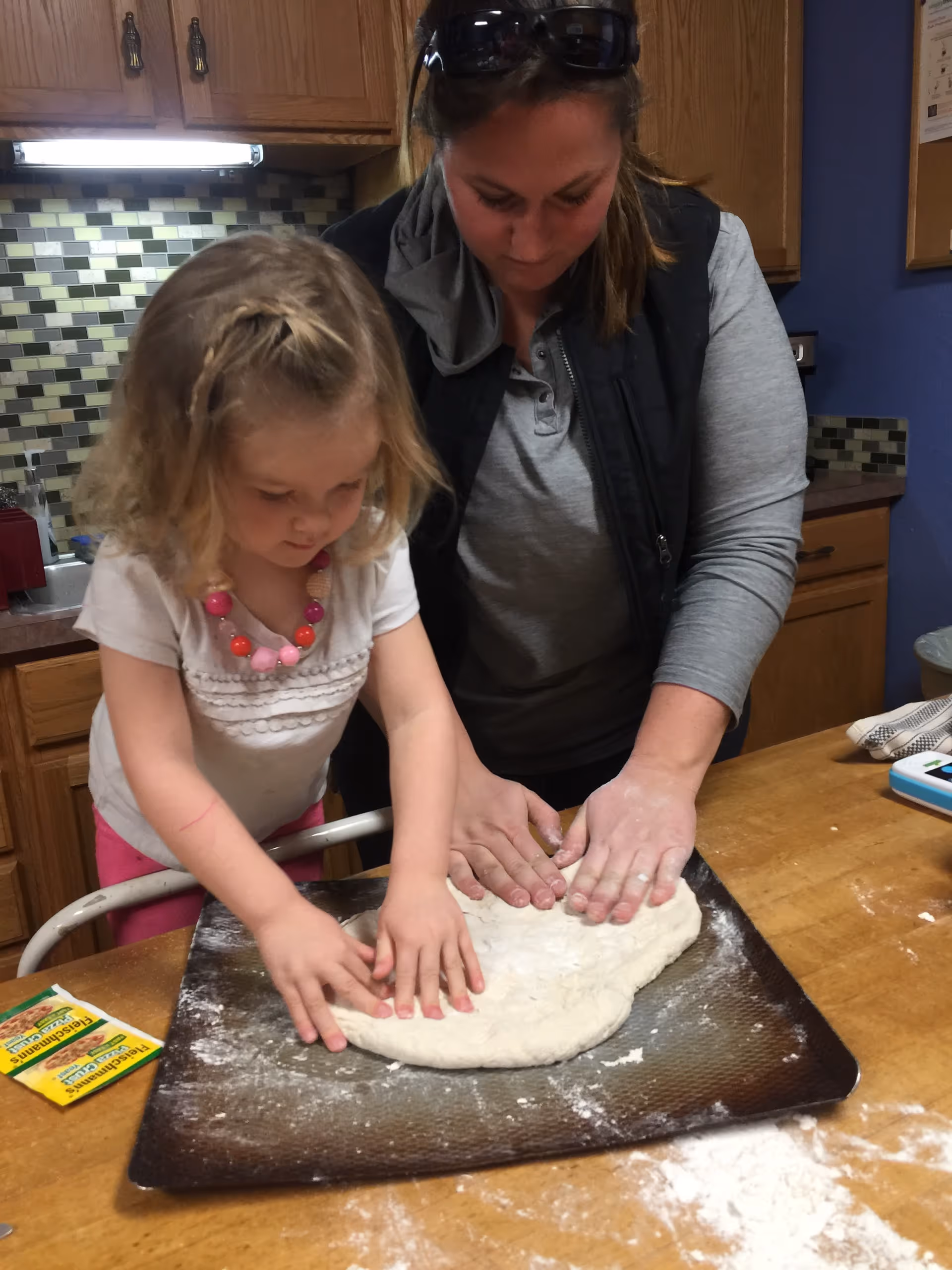 An adult woman and a young girl pressing and shaping dough together on a baking sheet in a kitchen.