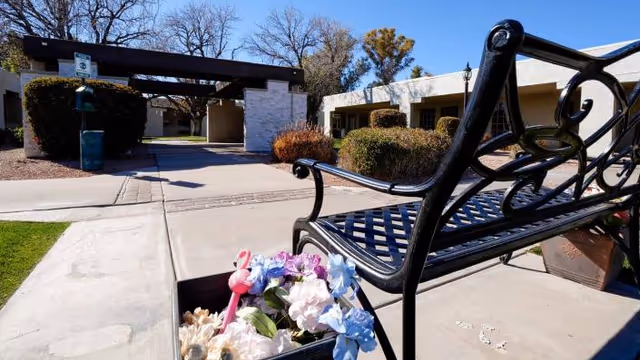 Outdoor seating area at Chandler Post Acute and Rehabilitation featuring a black metal bench next to a planter with colorful artificial flowers. In the background, there is a pathway leading to a building entrance surrounded by bushes and trees under a clear blue sky.