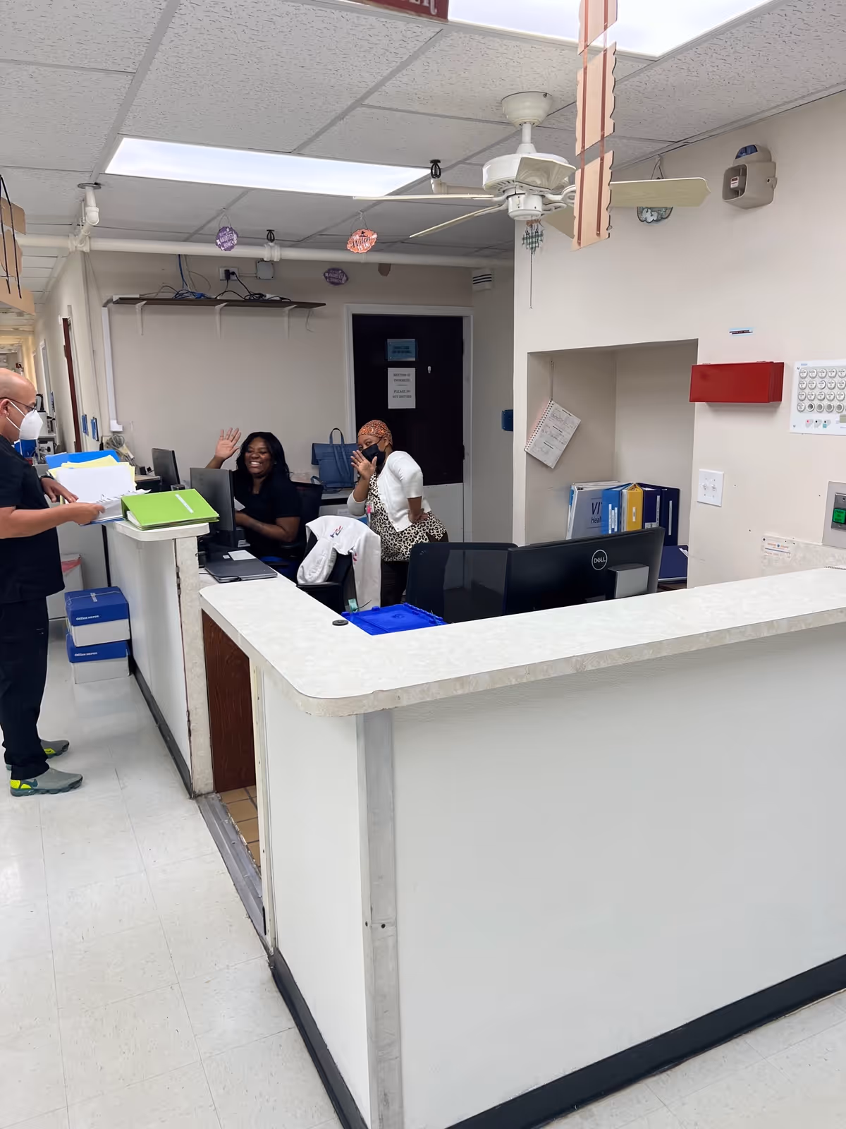 A nursing home reception or nurse station area with a white counter and two women sitting behind it, one waving and smiling, the other wearing a headscarf and a mask. A man wearing a mask is standing on the left side holding a folder. The room has white walls, a ceiling fan, fluorescent lighting, and office equipment such as computers and files.