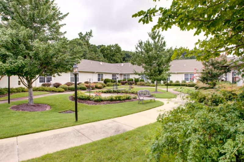 A landscaped outdoor garden area with a concrete walkway, green grass, trees, shrubs, and a bench. In the background, there is a single-story building with beige siding and multiple windows, surrounded by more greenery.