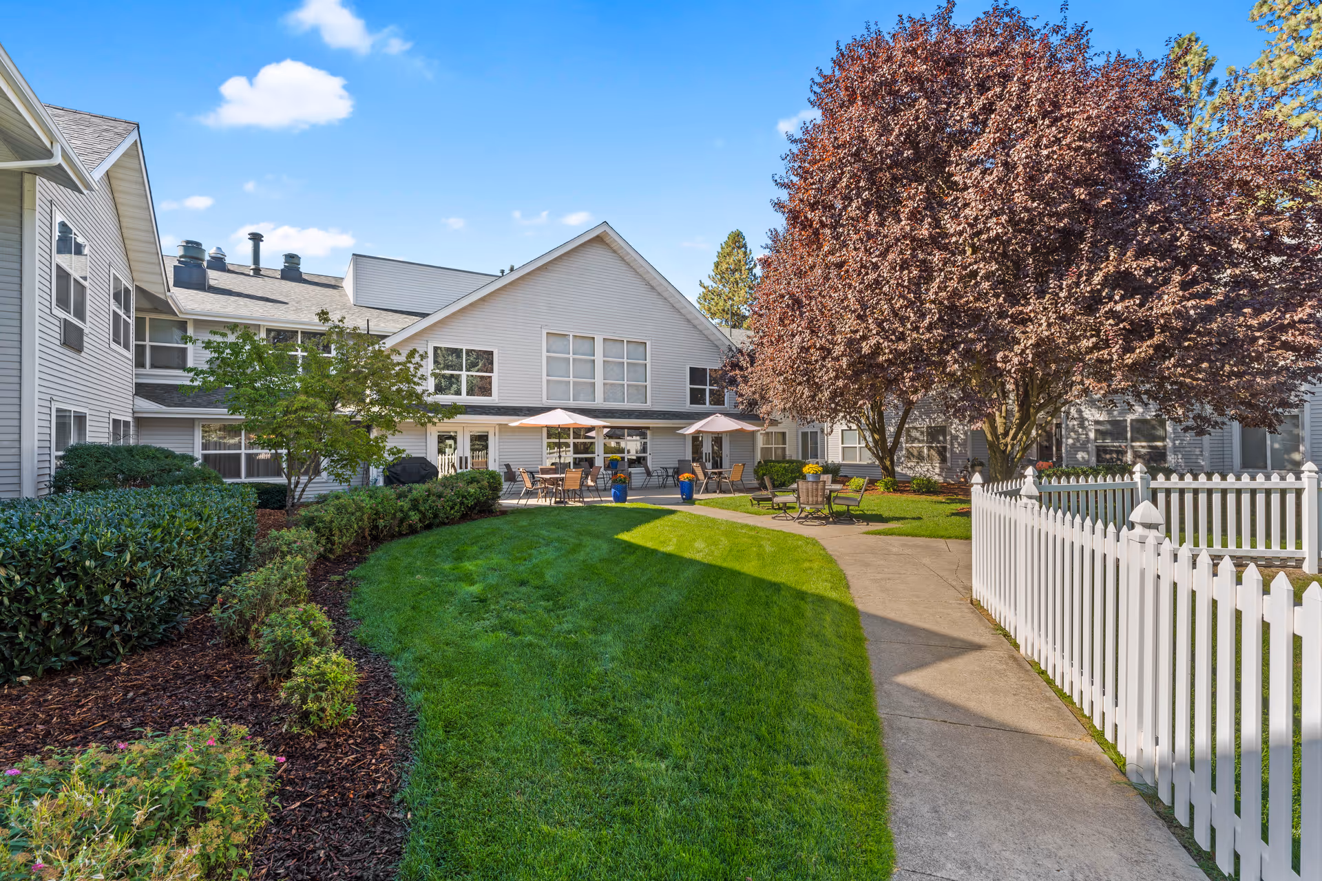 Outdoor courtyard area of a senior living facility with well-maintained green grass, a concrete walkway, a white picket fence, several trees including one with reddish leaves, and patio tables with umbrellas and chairs near the building.