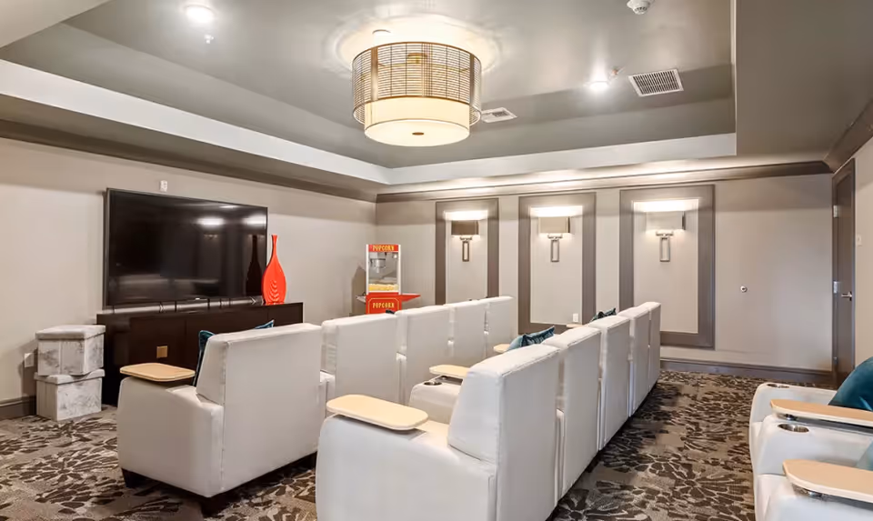 A cozy media room with two rows of white cushioned chairs facing a large flat-screen TV mounted on a dark wooden cabinet. There is a red decorative vase on the cabinet and a popcorn machine in the corner. The room features patterned carpet, soft wall sconces, and a modern ceiling light fixture.