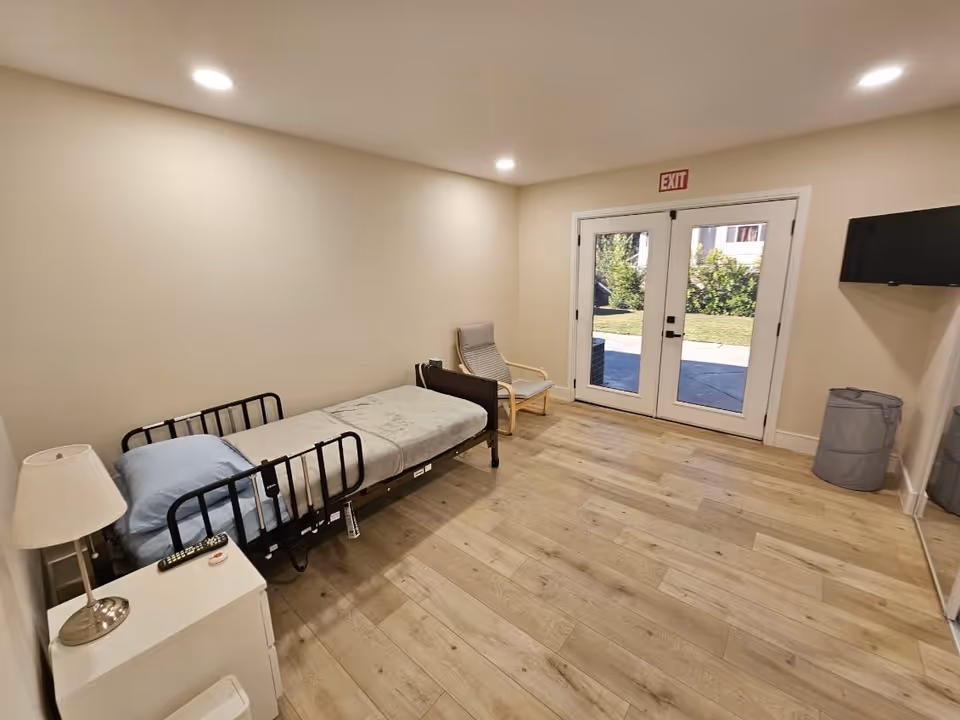 Simple bedroom with an adjustable single bed, bedside table and lamp, a chair, wall-mounted TV, and double glass doors opening to an outdoor area.