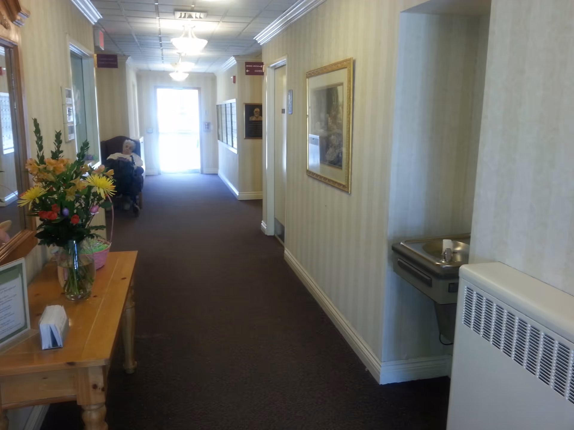 A hallway in a senior living facility with beige wallpaper and carpeted floor. On the left side, there is a wooden table with a vase of flowers and some papers. Further down the hallway, an elderly person is sitting in a wheelchair near the exit door. On the right side, there is a drinking fountain and a framed picture on the wall. Ceiling lights illuminate the corridor.