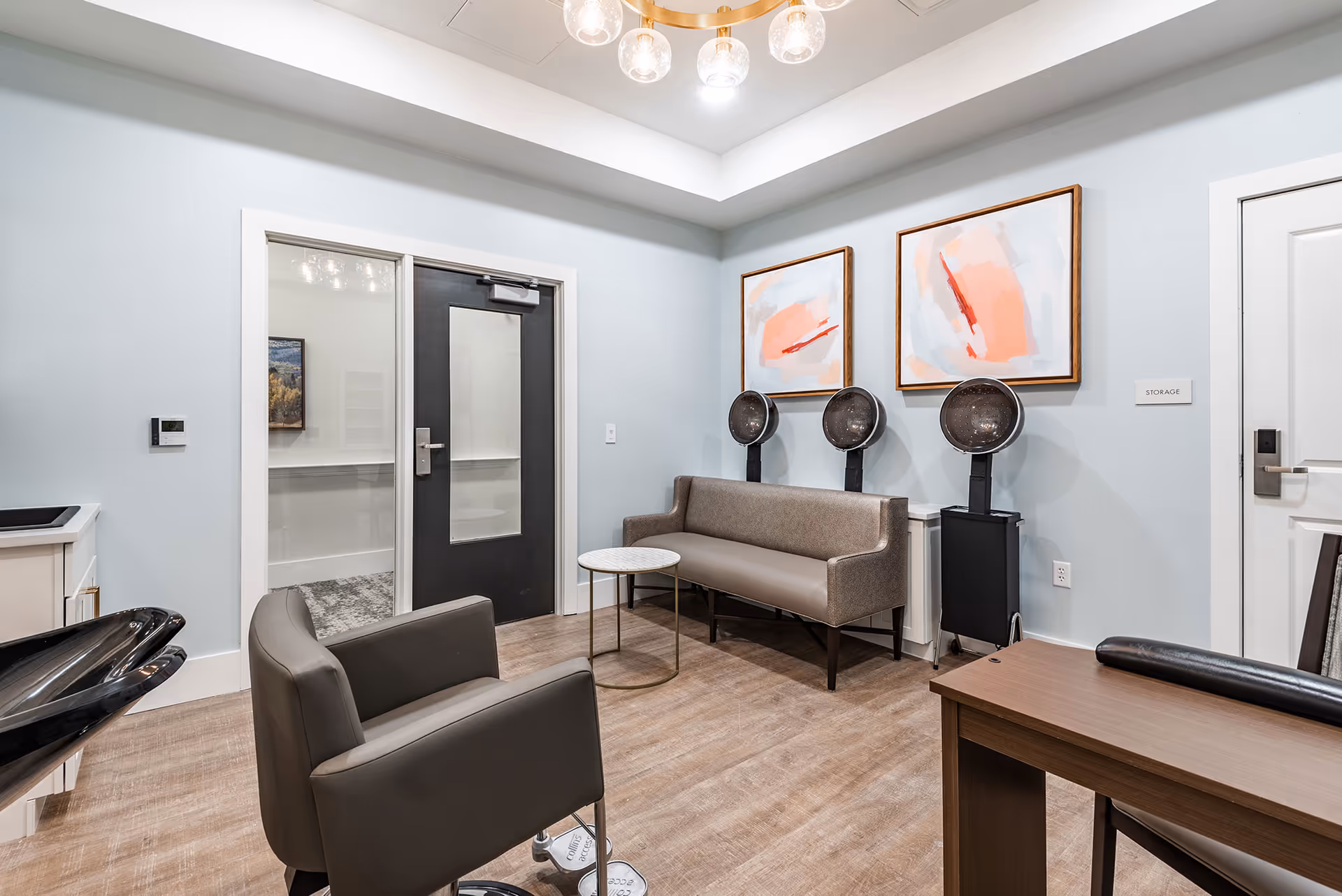 A modern salon room with three hair drying stations along the wall, a gray cushioned bench, a small round table, a gray salon chair, and a wooden desk. The walls are light blue with two abstract paintings hanging above the bench. There is a black door with a window and a white door labeled 'STORAGE'.