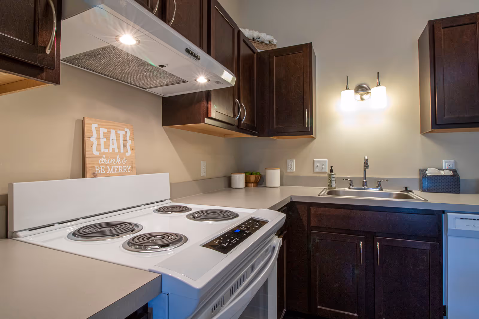 A kitchen corner featuring a white electric stove with four coil burners, dark wooden cabinets, a stainless steel sink with a faucet, a wall-mounted light fixture above the sink, and a decorative wooden sign that reads 'EAT drink & BE MERRY'. There are also small containers and a soap dispenser on the countertop.