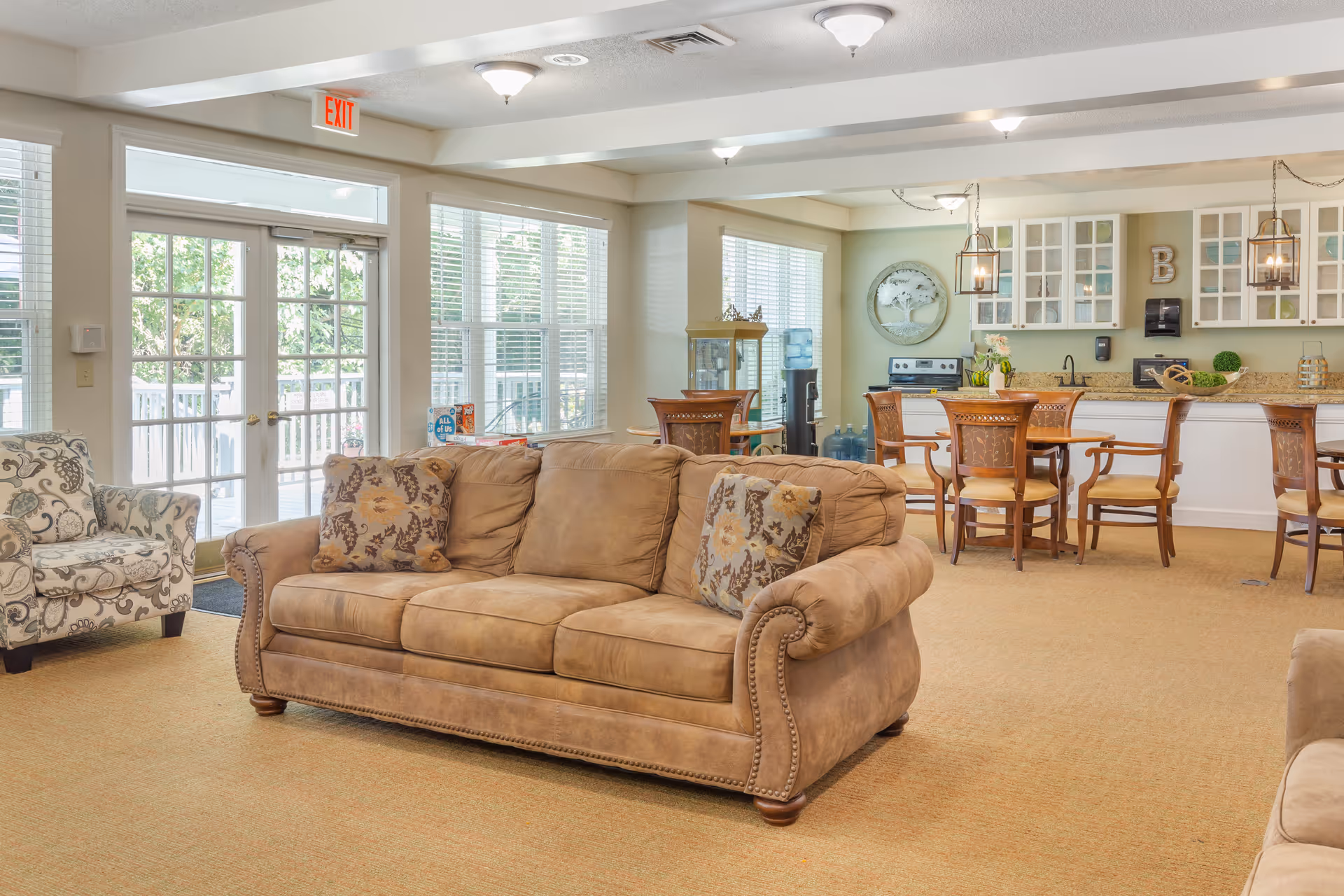 A bright and spacious common area in a senior living facility featuring a tan sofa with patterned cushions, a floral armchair, and a dining area with wooden chairs and tables. The room has large windows with white blinds, a glass door leading outside, and a kitchen area with white cabinets and granite countertops in the background.