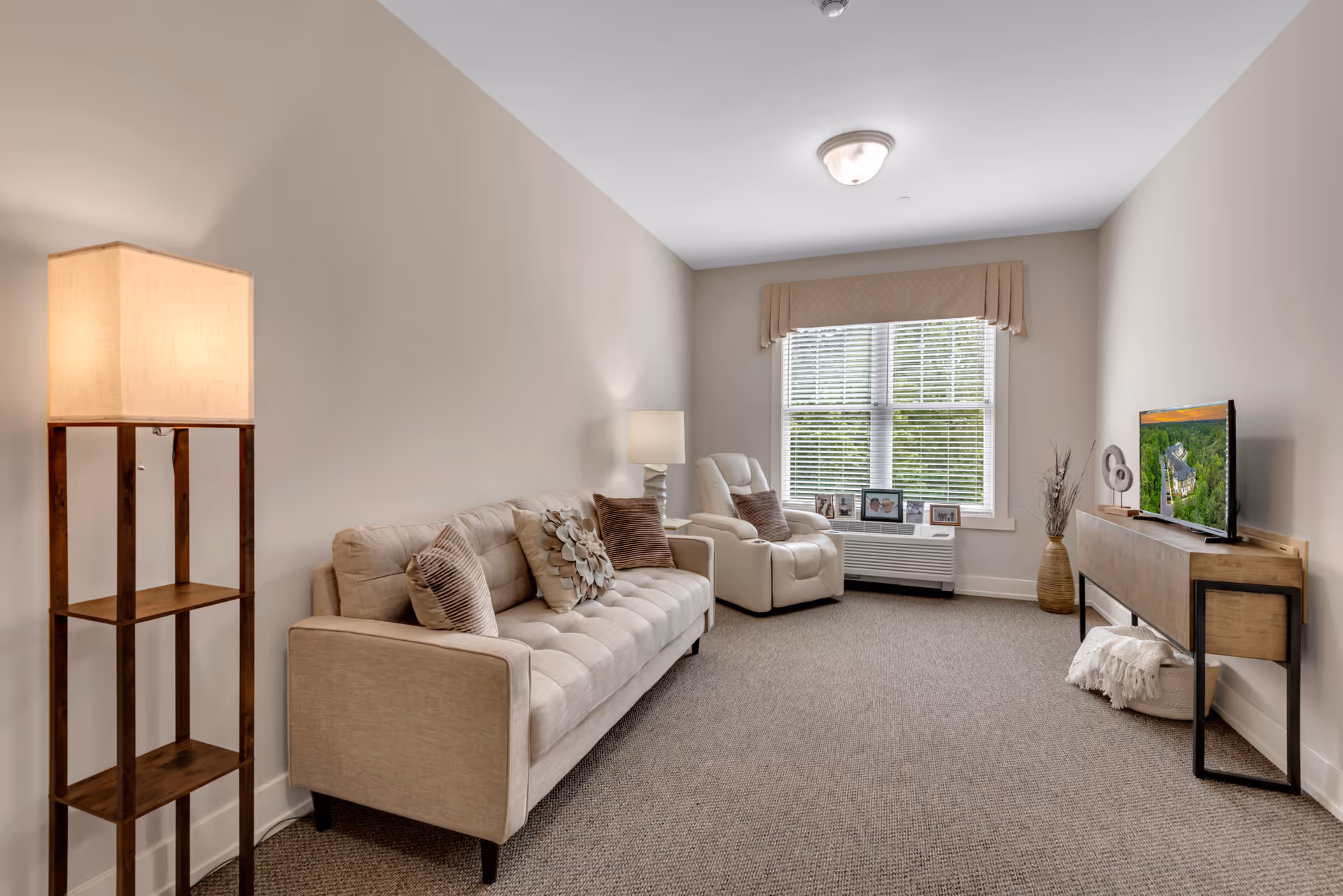 A cozy living room with a beige sofa adorned with decorative pillows, a white recliner chair, a wooden floor lamp with a square shade, a wooden TV stand with a flat-screen TV, and a window with blinds and a beige valance letting in natural light.